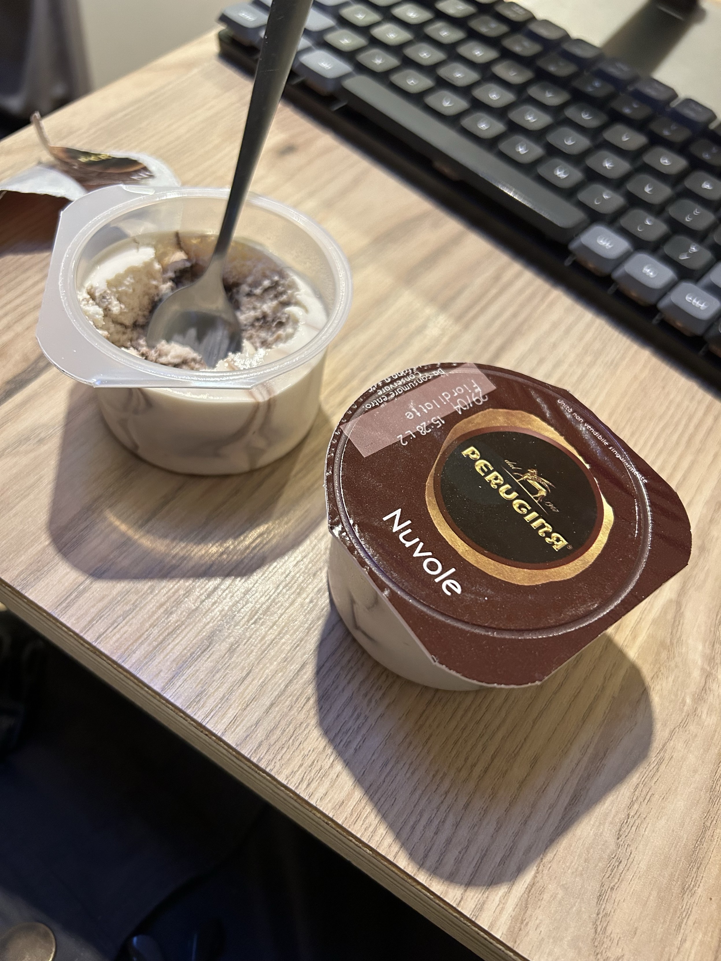 Otwarte i nieotwarte pojemniki z deserem z łyżeczką w jednym, na drewnianej powierzchni obok klawiatury komputera.