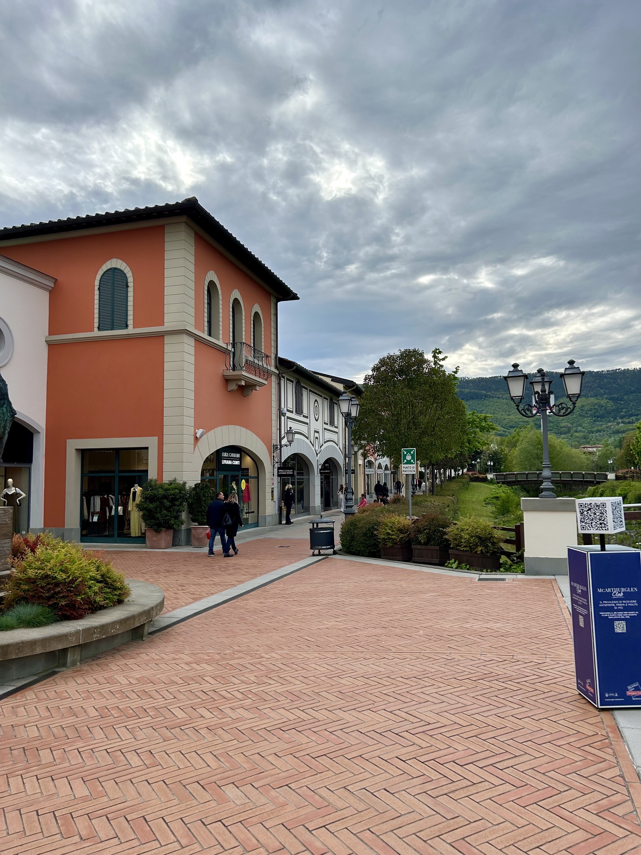 Zewnętrzny plac centrum handlowego z ceglanym chodnikiem, spacerującymi ludźmi i ozdobnymi latarniami, na tle zachmurzonego nieba i zielonych wzgórz.