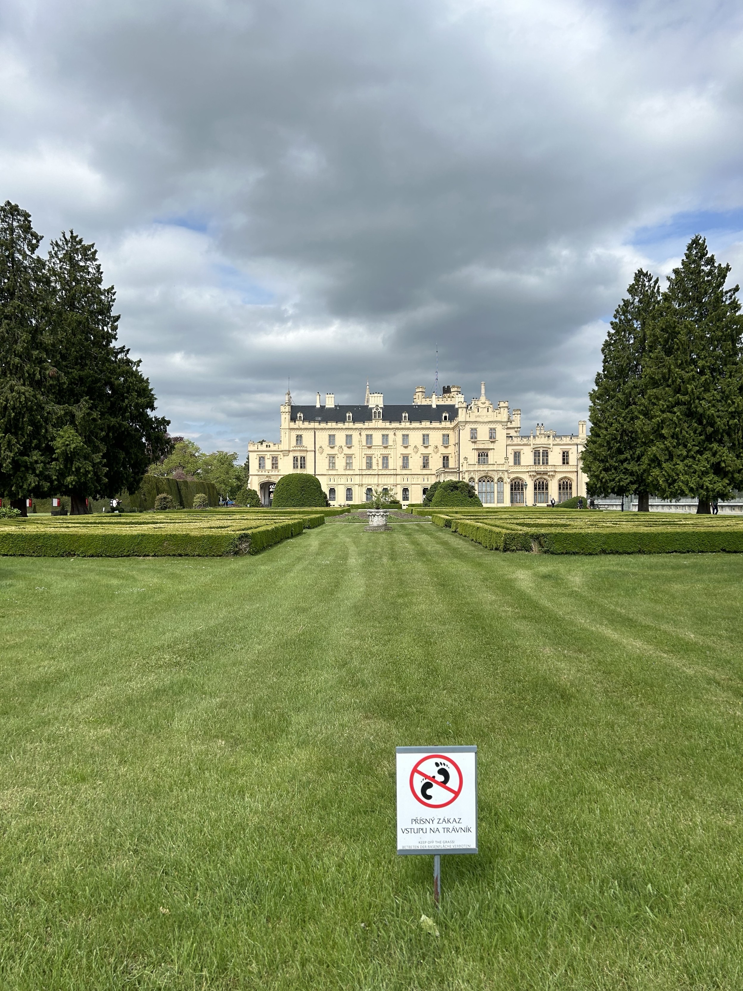 Duży pałac widziany przez zadbany trawnik z zachmurzonym niebem powyżej i znakiem na pierwszym planie zakazującym chodzenia po trawie.